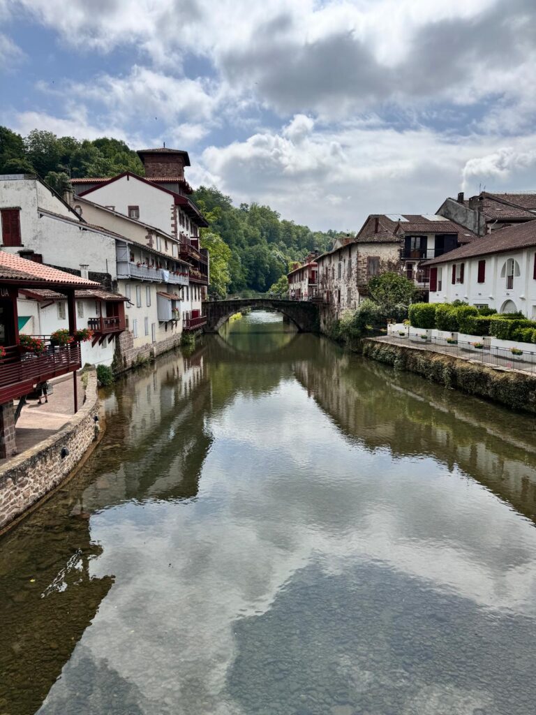 A scenic view of a stone bridge and Basque-style houses along a calm river in Saint-Jean-Pied-de-Port, France, with green hills and a cloudy sky in the background.