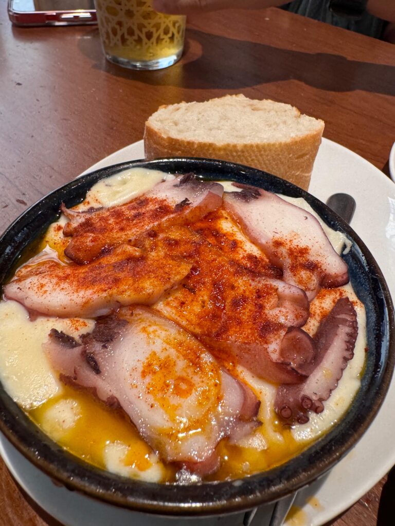 A bowl of Pulpo a la Gallega with octopus, melted cheese, and paprika, served with a slice of bread on a wooden table.