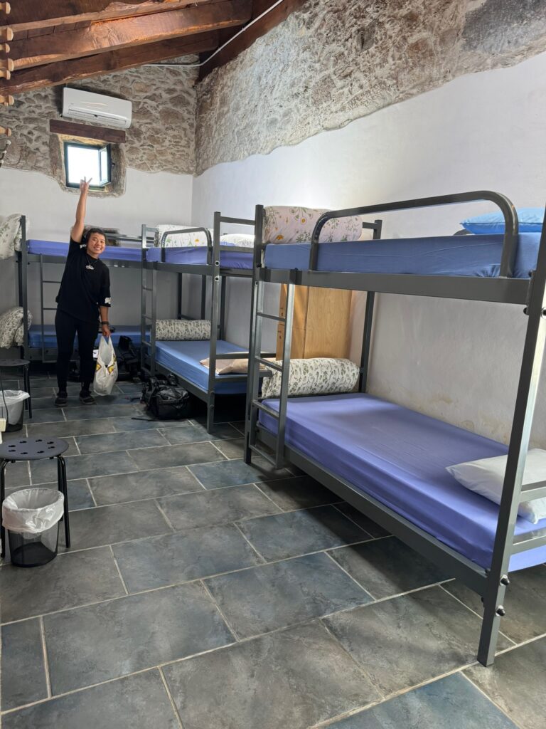 Controversial Topic: Team Book Ahead or Team Wing it - A cheerful female hiker in a tidy bunk room with stone floors, eagerly preparing to wash her laundry after a long day on the Camino.