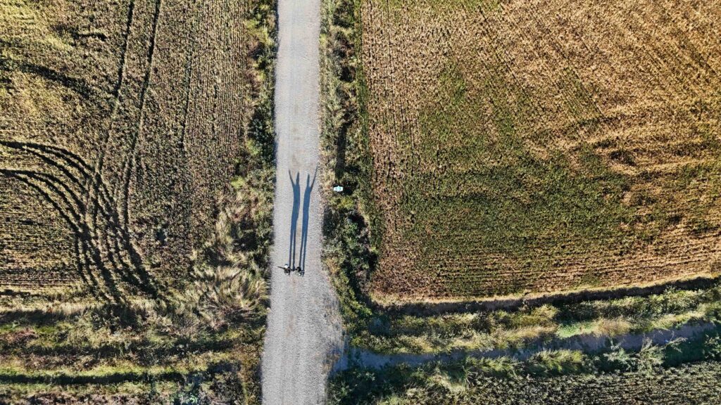 Aerial shot of two hikers on a dirt road along the Camino de Santiago, with green and dried fields on each side and shadows stretching across the road.