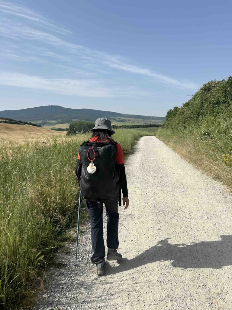 A tired hiker walking along a straight road with a Camino shell hanging from her backpack, symbolizing the pilgrimage.