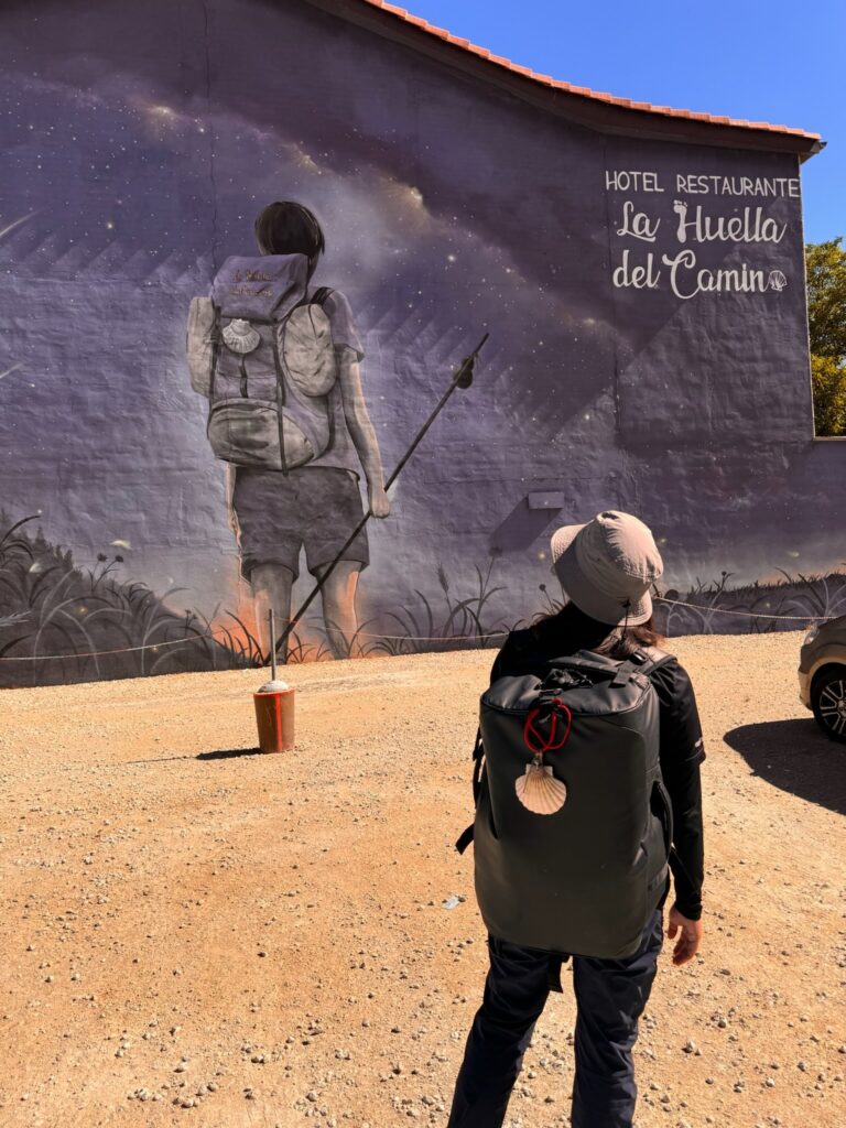 A pilgrim with a backpack and a Camino shell stops to admire a mural showing another pilgrim under a starry night, carrying the same symbol.