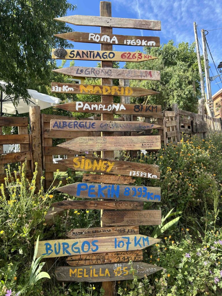 A wooden signpost along the Camino de Santiago pointing to Roma, Santiago, Sydney, and Burgos, with distances marked.