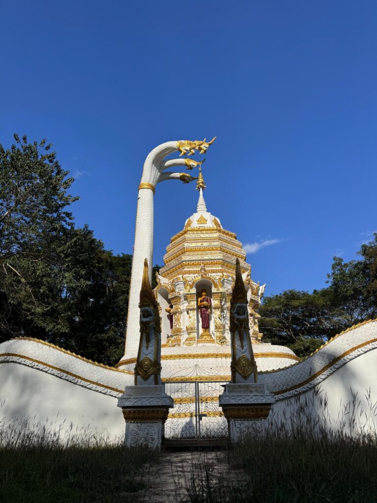 Ornate white and gold stupa with naga sculptures at a temple near Chiang Dao Caves in northern Thailand, set against a clear blue sky and surrounded by trees.