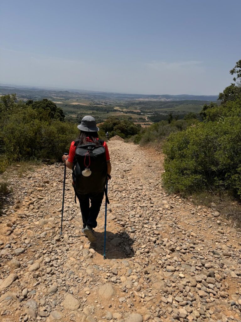 Pilgrim walking downhill on a rocky path after Alto de Perdón on the Camino de Santiago, with wide views of the Spanish countryside