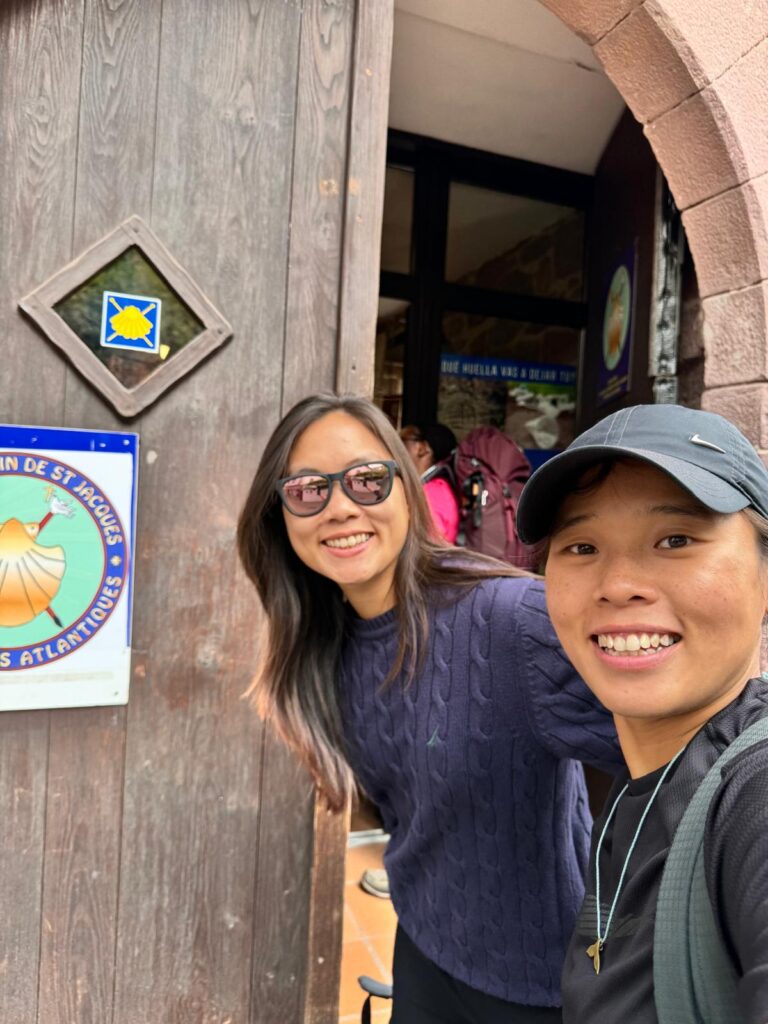 Two pilgrims smiling outside the Pilgrim Office in Saint-Jean-Pied-de-Port, holding their Camino documents and scallop shells.